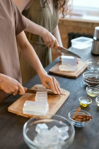 Hands of two young women cutting hard soap mass into cubes on boards by table