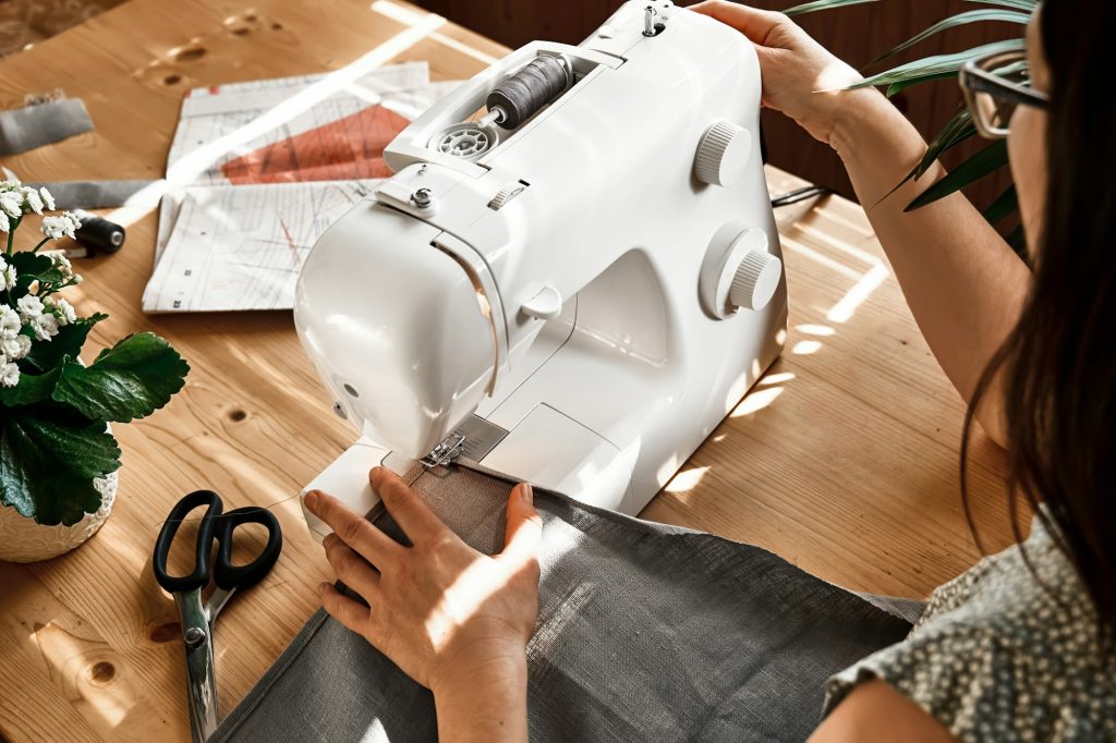 Woman sewing on sewing machine in small studio. Fashion atelier, tailor craft, handmade clothes.